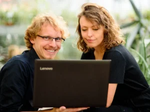 Projekt ogrodu - cena. Na zdjęciu uśmiechnięci projektanci przed laptopem. W tle zieleń. 
